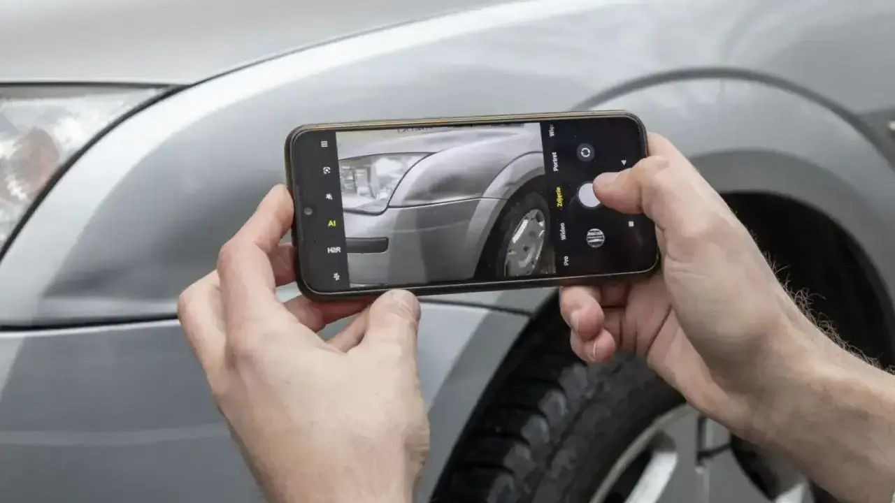 Ręce trzymają smartfon, fotografując reflektor i błotnik srebrnego auta. Sprawdzam lakierowanie elementu samochodu cennik.