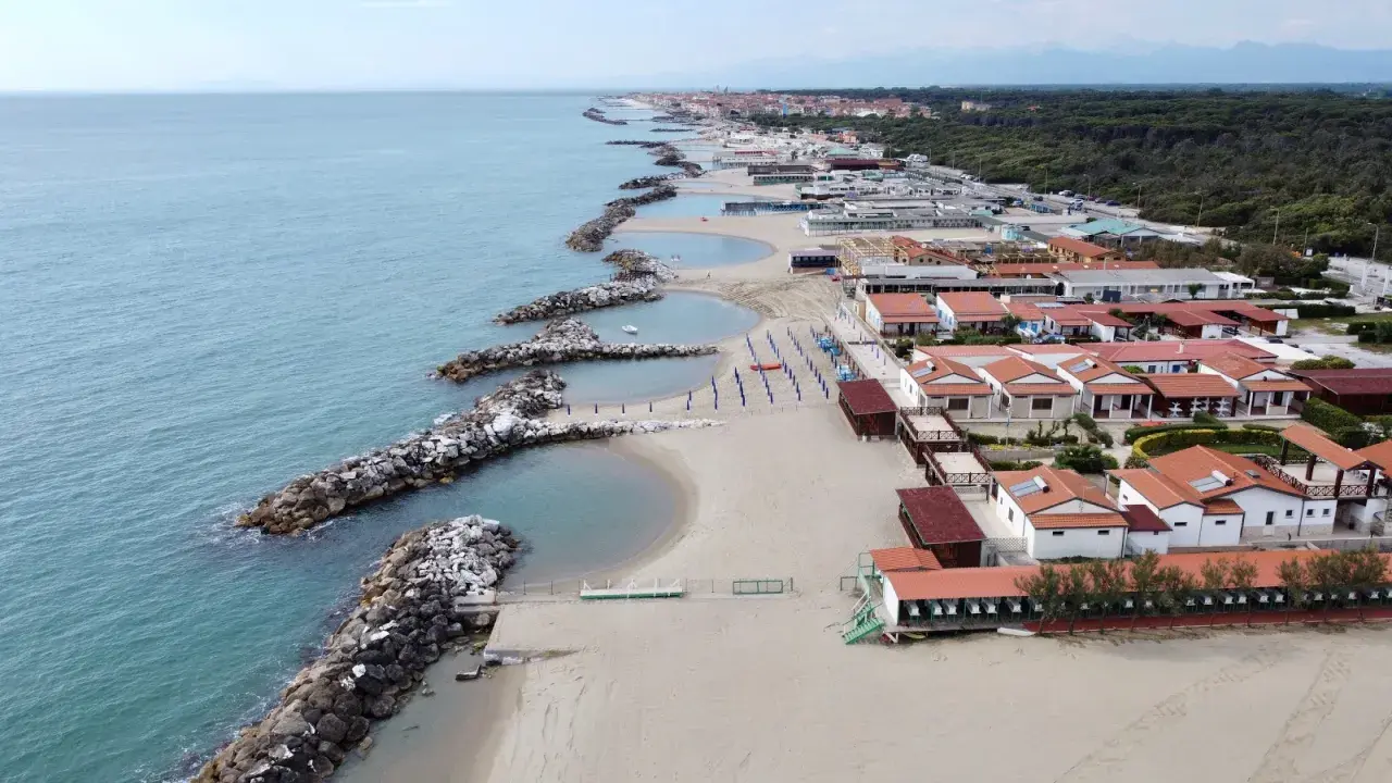 Vacker vy över pisa strand med sina stenpirar som bryter vågorna. Längre bort syns hus och en grönskande skog.