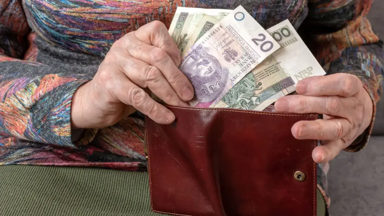 Dłoń starszej osoby wkłada polskie banknoty do brązowego portfela. Widać nominały 20, 100 i 50 złotych, stanowiące ekwiwalent codziennych wydatków.