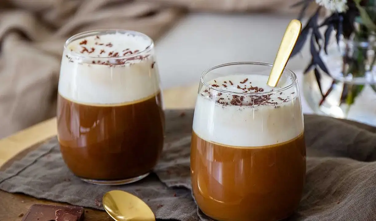 Två glas med krämig kaffe Karlsson, toppad med vispgrädde och chokladströssel. En gyllene sked vilar i det ena glaset.