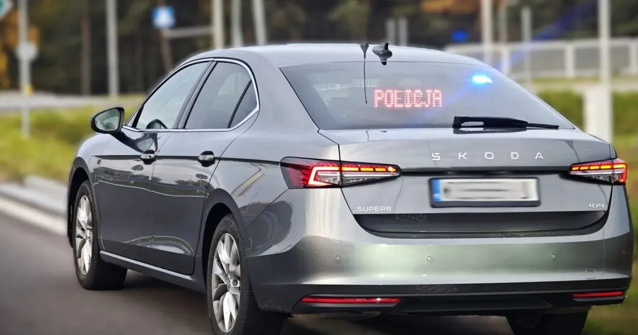 Szary, nieoznakowany radiowóz Skoda Superb z włączonymi niebieskimi światłami i napisem "POLICJA" na tylnej szybie.