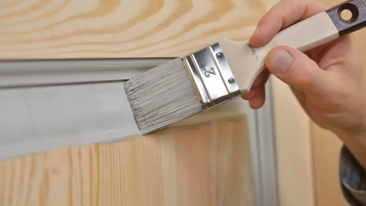 Main d'une personne peignant une porte en bois sans la dégonder, appliquant une peinture grise avec un pinceau.