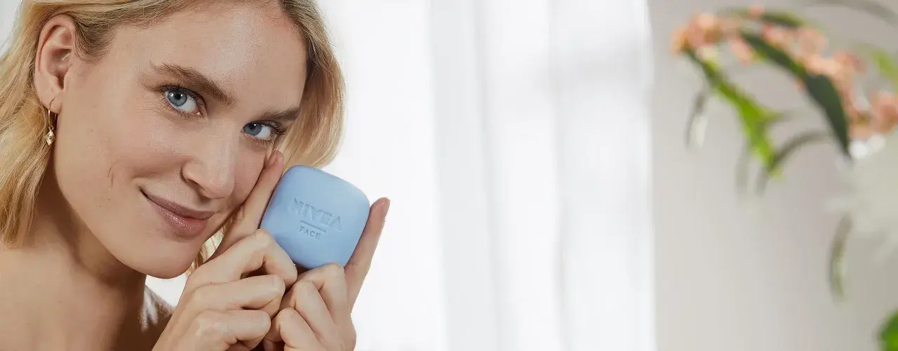Une femme blonde tient un pain de savon bleu Nivea Face. Elle sourit, sa peau semble douce. Ce savon a un pH neutre, combien est-il doux pour votre visage ?