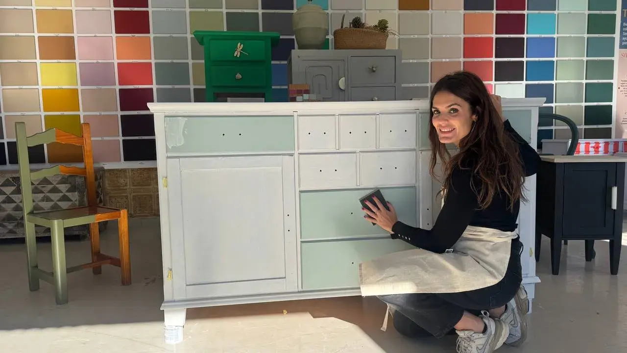 Mujer sonríe mientras trabaja en un mueble, aplicando pintura blanca. Aprende como pintar un mueble oscuro en blanco sin lijar.