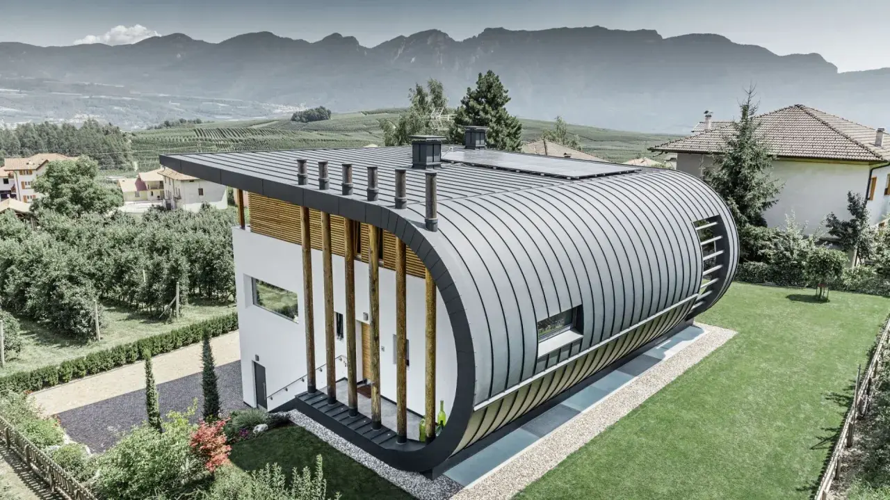 Nowoczesny dom z zaokrąglonym dachem z aluminium, które nie koroduje, otoczony zielenią i górami.