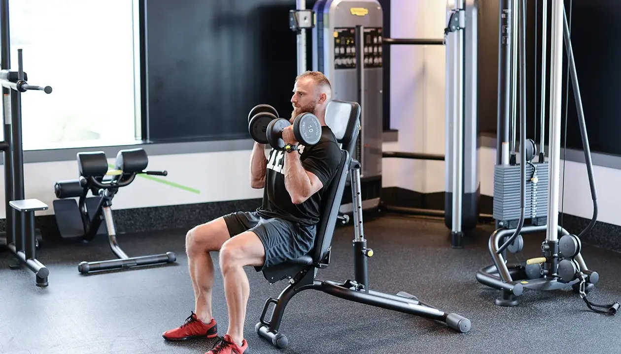 Mężczyzna wykonuje ćwiczenie z hantlami na ławce. Arnoldki stojąc, to nie jego cel, skupia się na sile.