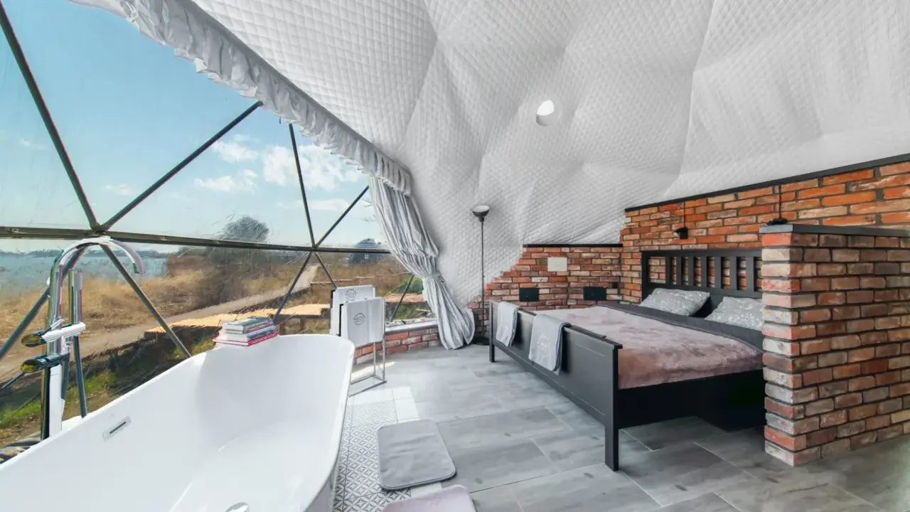 Luksusowy glamping nad jeziorem: wanna z widokiem, łóżko i ceglana ściana w kopułowym wnętrzu.