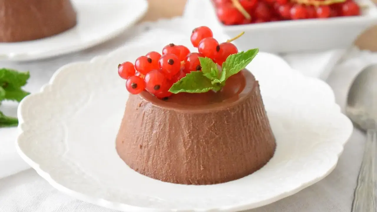 Panna cotta czekoladowa z czerwonymi porzeczkami i miętą na białym talerzyku.