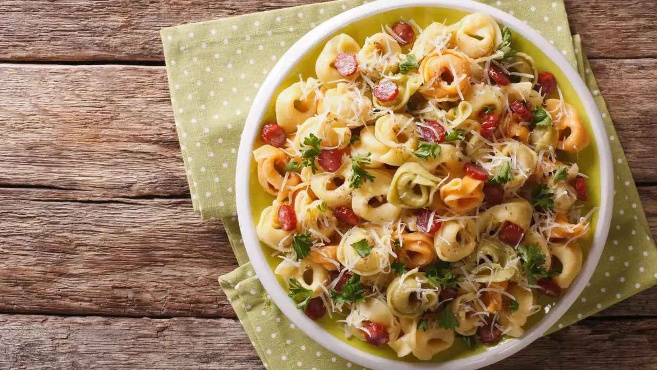 Kolorowa sałatka z tortellini, kawałkami kiełbasy i parmezanem, udekorowana natką pietruszki, podana na talerzu na zielonej serwetce w kropki.