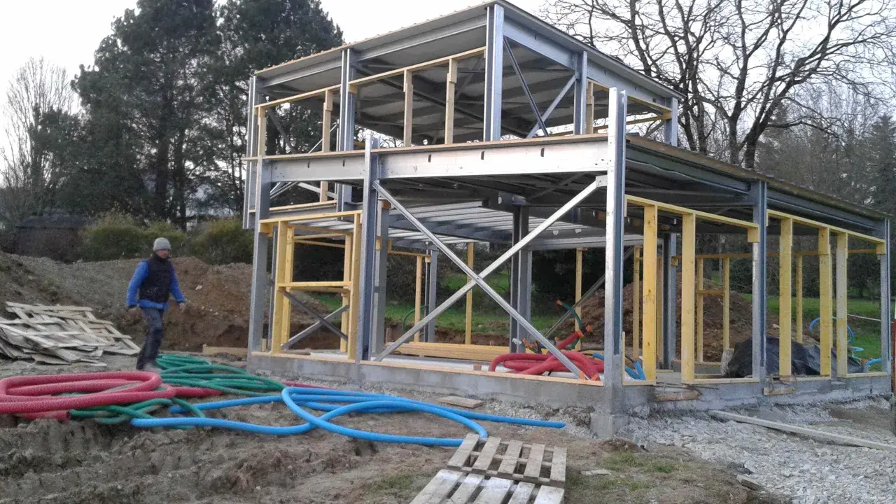 Chantier d'une maison ossature métallique, structure en acier et bois, avec des tuyaux colorés au sol.