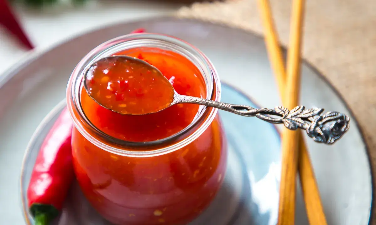 Ein Löffel mit leuchtend roter Thai Chilli Sauce, daneben frische Chilis und Stäbchen.