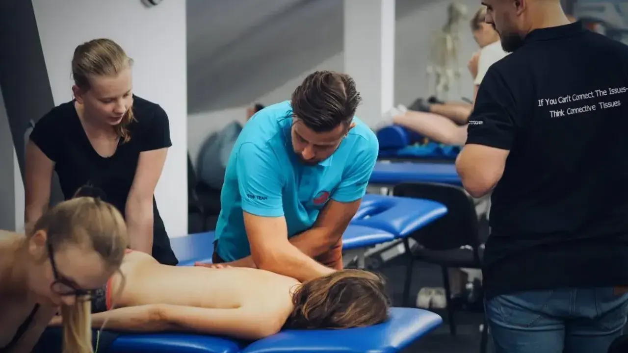 Uczestnicy szkolenia wykonują masaż tkanek miękkich na pacjencie leżącym na kozetce. Terapeuta w niebieskiej koszulce demonstruje technikę.