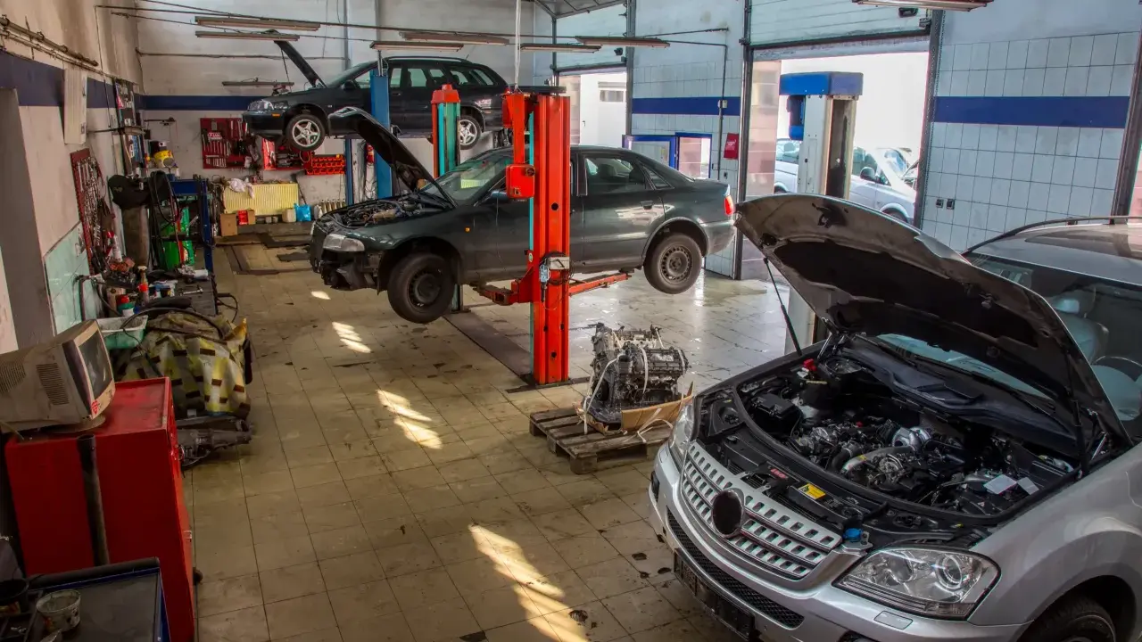 Warsztat samochodowy z kilkoma autami na podnośnikach. Widać m.in. ciemny sedan, który wygląda jak p1351 peugeot, oraz srebrny SUV z otwartą maską.
