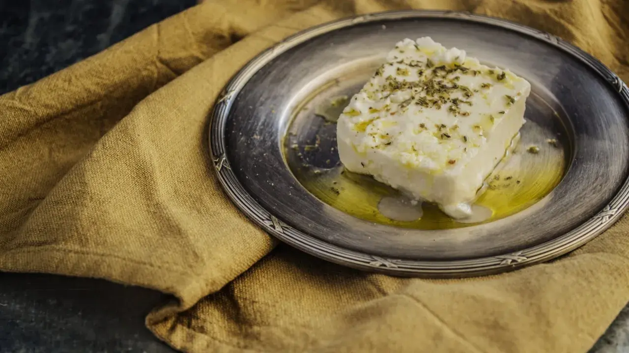 Ein Stück cremiger, vegetarischer Feta mit Kräutern und Olivenöl auf einem silbernen Teller, daneben ein rustikales Leinentuch.