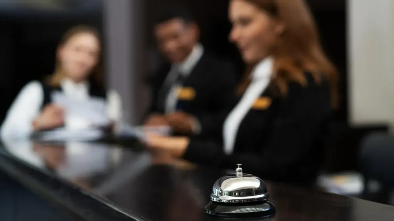 Dzwonek hotelowy na ladzie, za nim recepcjoniści pracują. Wzór karty meldunkowej w hotelu.
