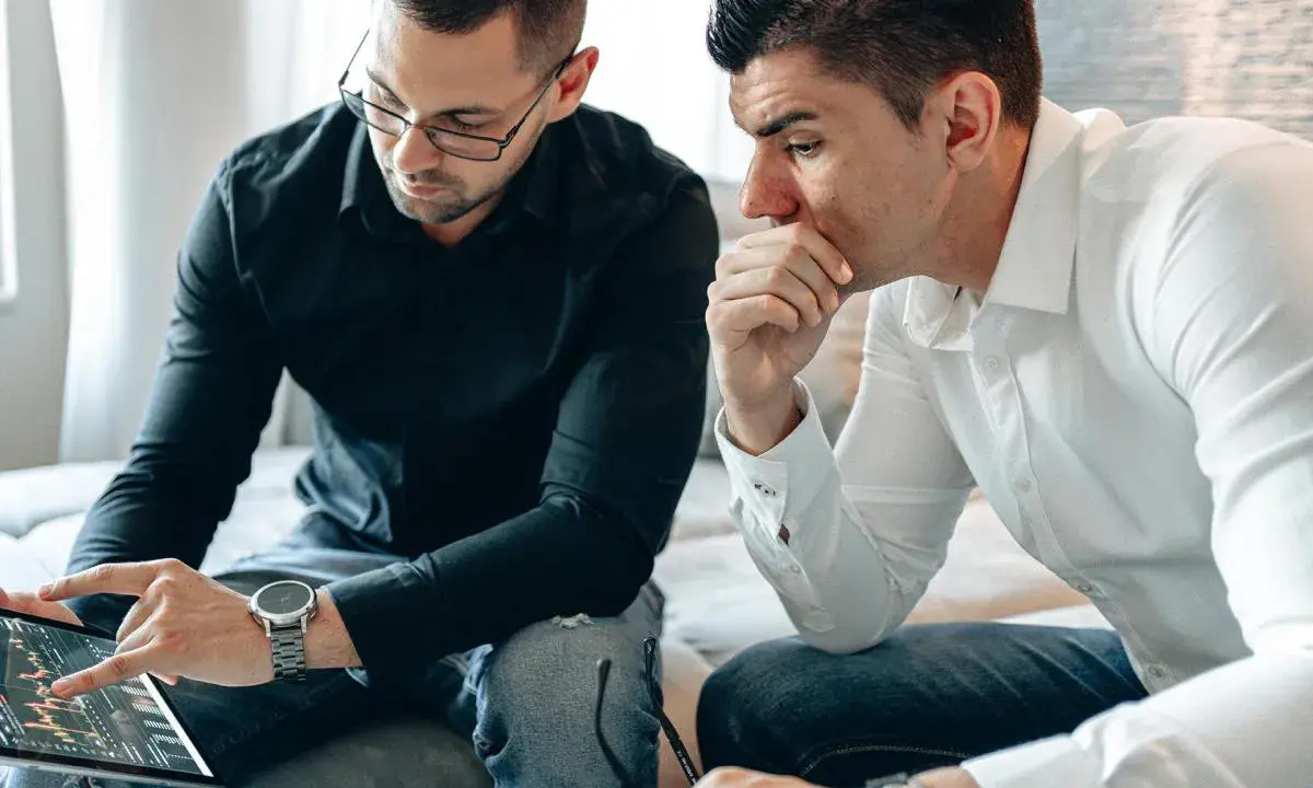 Dos hombres analizan gráficos en una tablet. Uno señala la pantalla, discutiendo cómo domiciliar la nómina para optimizar finanzas.
