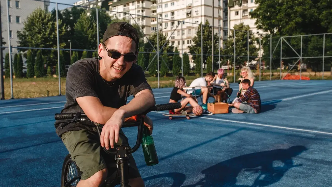 Uśmiechnięty chłopak na rowerze z butelką piwa, obok grupa znajomych. Może to być scena, która zakończy się mandatem za jazdę rowerem po alkoholu.