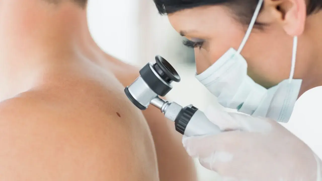 Lekarz w masce i rękawiczkach wykonuje dermatoskopię znamienia na ramieniu pacjenta.