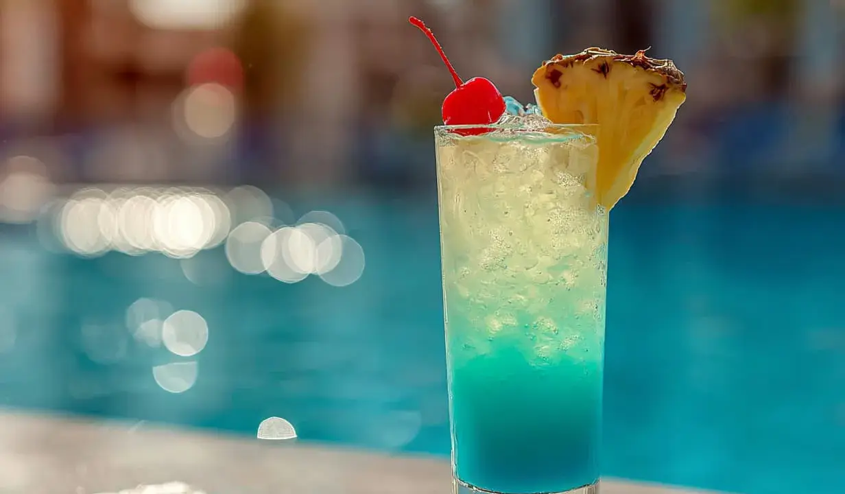En blå och gul drink med körsbär och ananas står vid en pool. Perfekt för en solig dag.