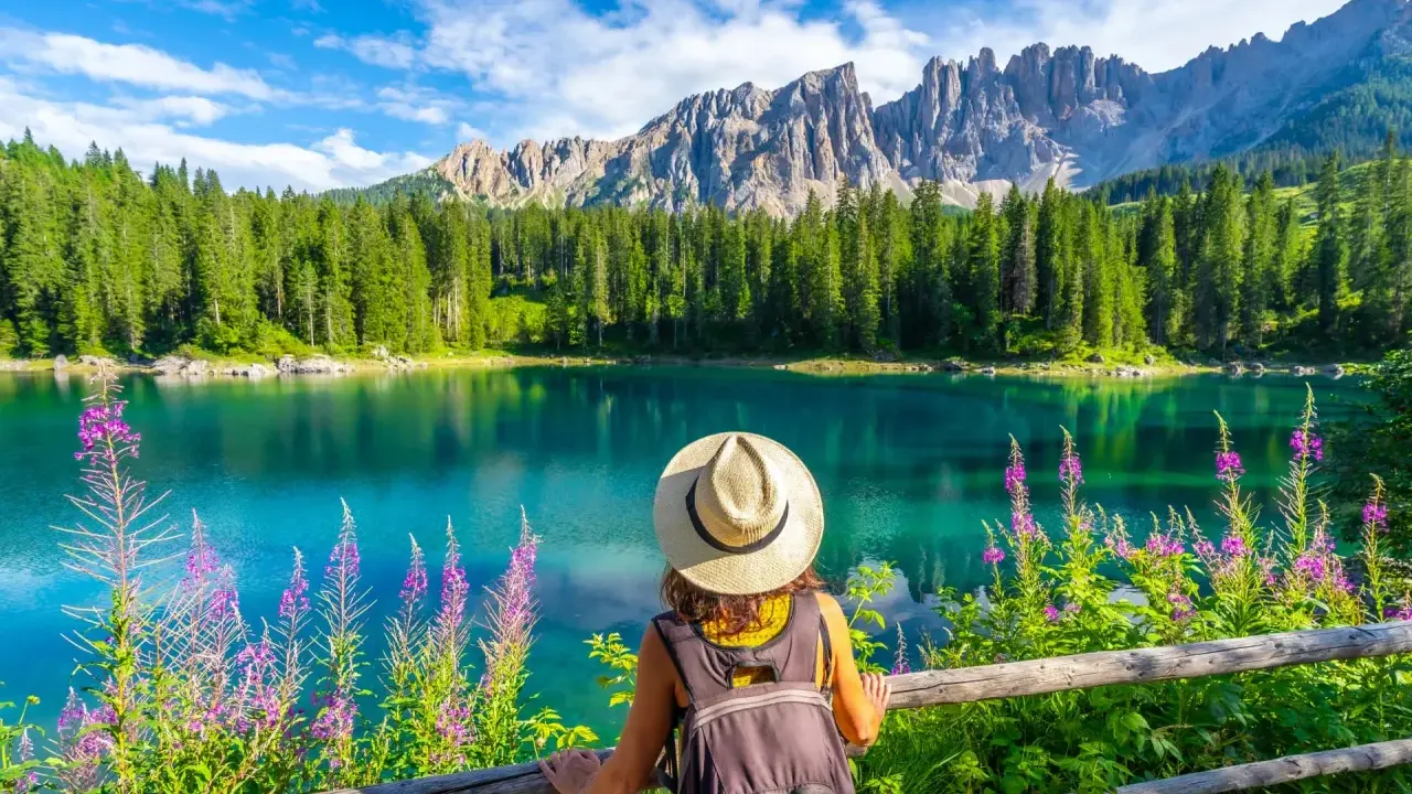 En kvinna med hatt och ryggsäck njuter av den vackra **Dolomiterna sommar**-utsikten över en turkos sjö, omgiven av skog och berg.