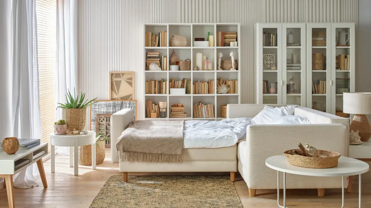 Jasny salon z sypialnią na małej powierzchni. Rozkładana sofa, regały z książkami i dekoracje tworzą przytulną przestrzeń.
