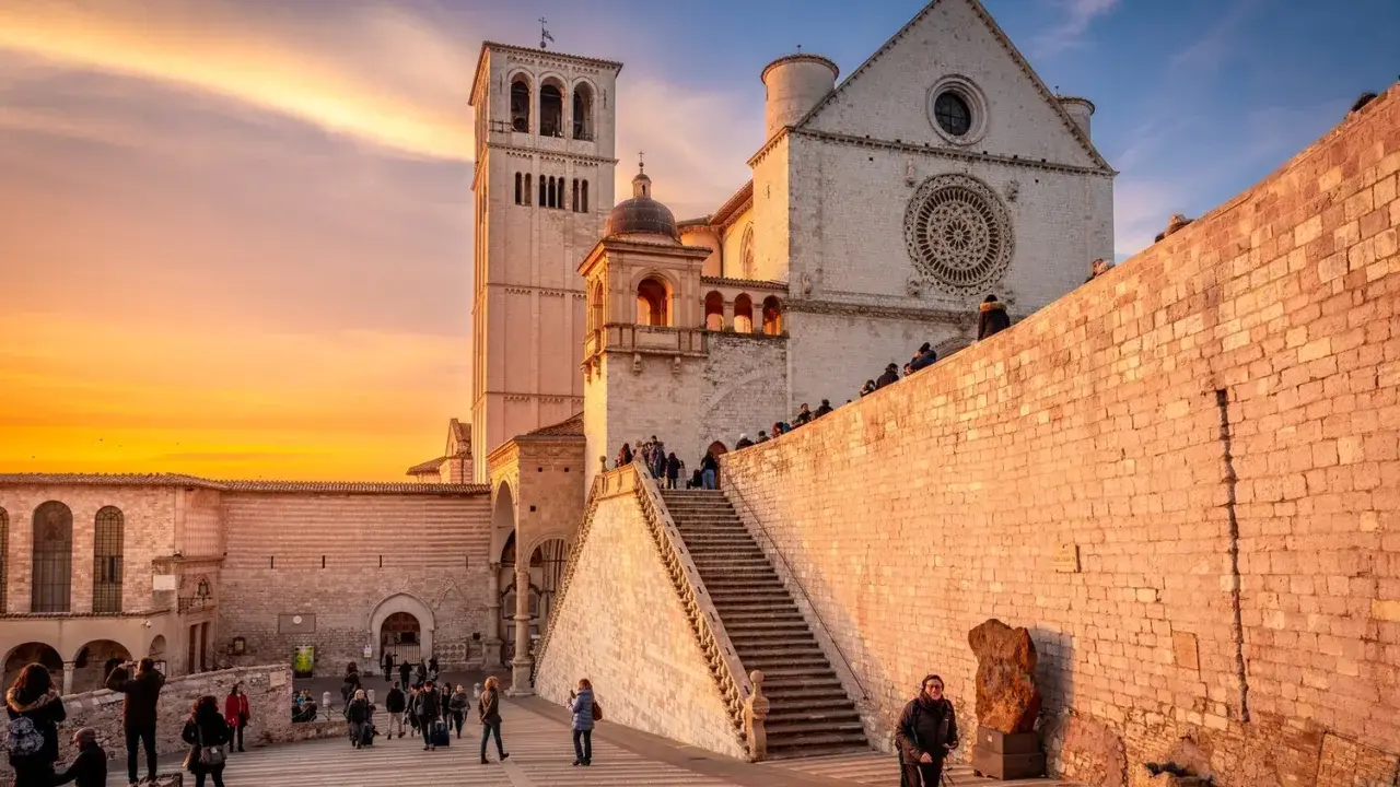 Assisi cosa vedere: la Basilica di San Francesco al tramonto, con la sua imponente facciata e la torre campanaria, illuminata da una calda luce dorata.