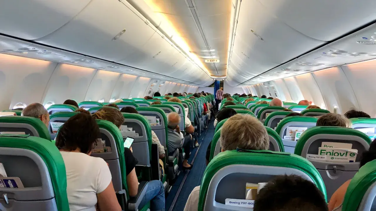 Wnętrze samolotu Transavia, pasażerowie siedzą w rzędach, przygotowani do lotu. Bagaż podręczny schowany w schowkach nad głowami.