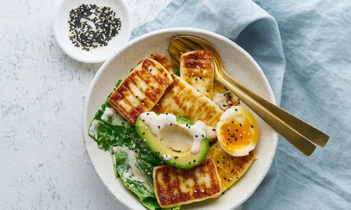 Szybki keto obiad: grillowany ser halloumi, awokado, jajko na miękko i zielone liście, posypane sezamem.