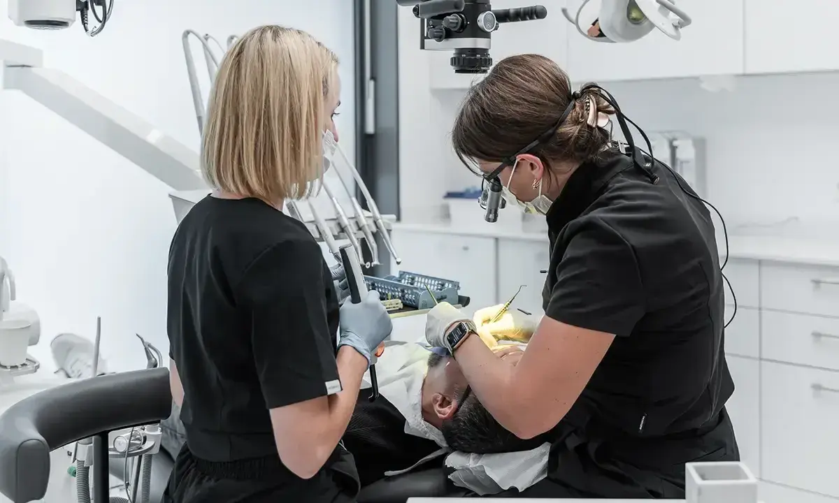 Dwie dentystki pracują przy pacjencie. Jedna używa mikroskopu, druga trzyma narzędzie. To obrazek pokazujący, jak zostać dentystą.