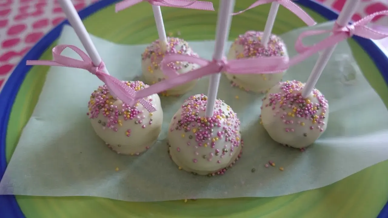 Sju söta cake pops med vit choklad och rosa, gula och lila strössel, dekorerade med rosa band.