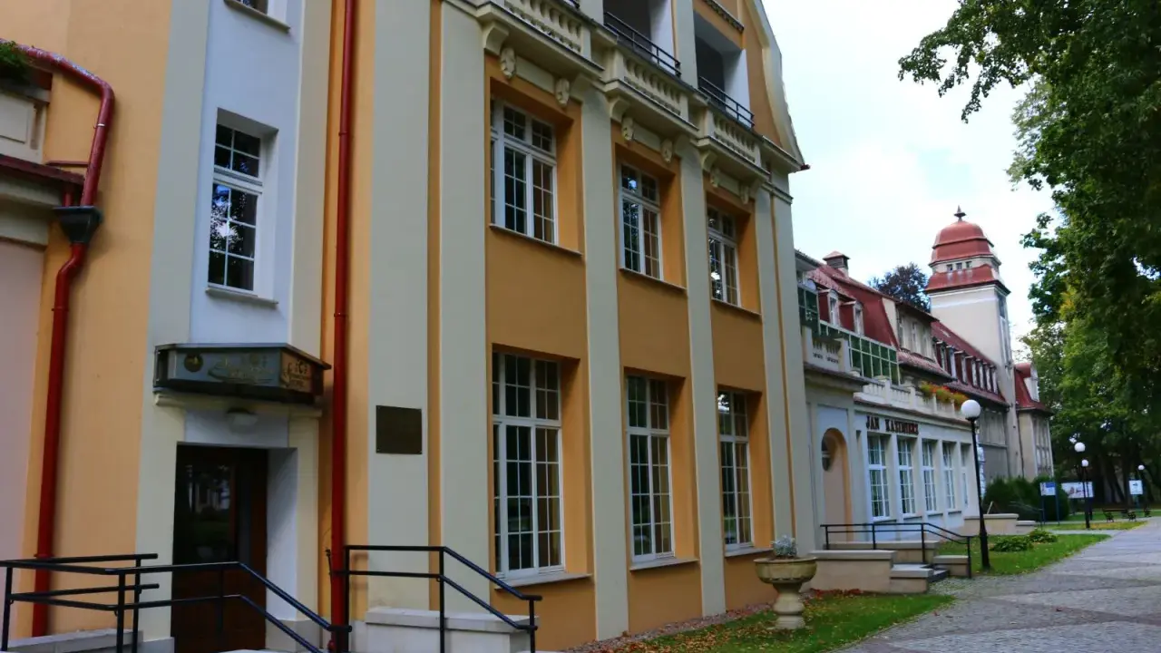 Sanatorium Jan Kazimierz w Dusznikach-Zdroju, z charakterystyczną żółto-kremową fasadą i czerwonym dachem, otoczone zielenią.