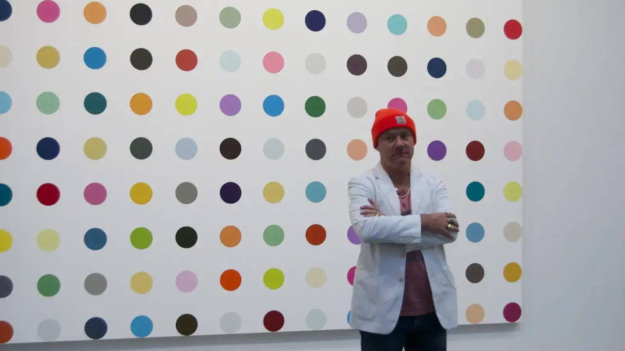 Damien Hirst steht vor seiner berühmten Punkt-Malerei, gekleidet in eine orangefarbene Mütze und einen weißen Blazer.