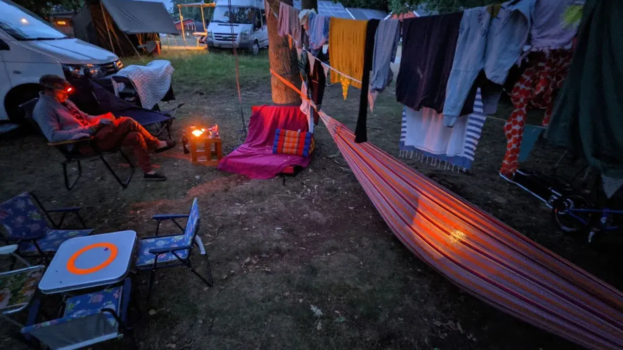 Una persona lee en una silla junto a una fogata, ropa secándose al aire y una hamaca iluminada. Disfrutando de una tranquila acampada al aire libre.