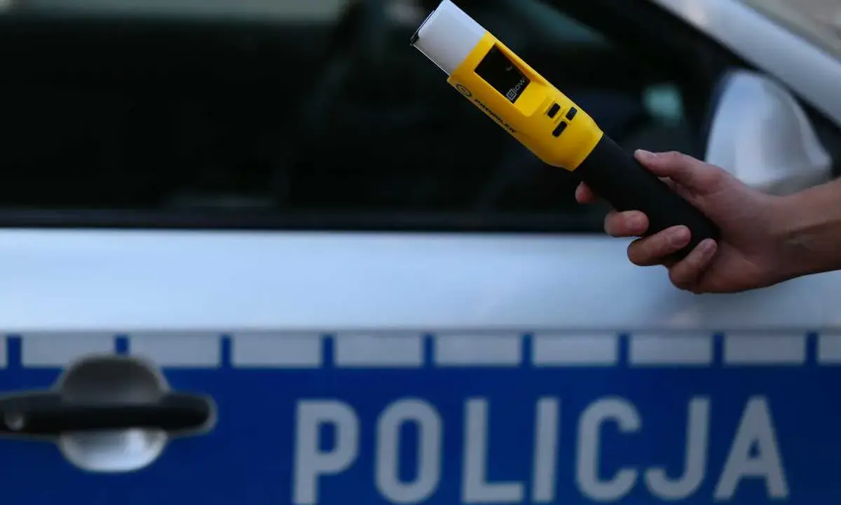 Policjant przeprowadza badanie trzeźwości nieletniego przy użyciu alkomatu. Na samochodzie widnieje napis "POLICJA".