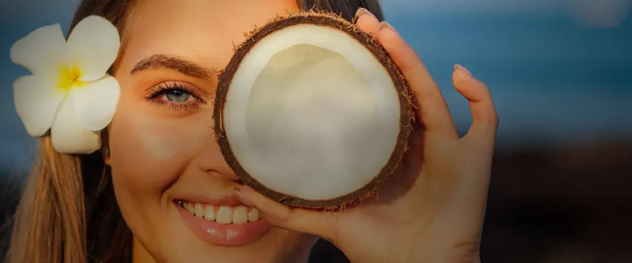 Donna sorride con un fiore tra i capelli, coprendo un occhio con una noce di cocco. Attenzione: olio di cocco sul viso fa male.