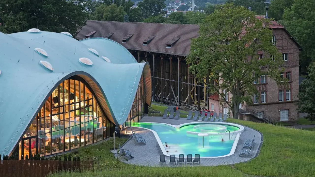 Moderne Therme mit geschwungenem Dach und beleuchtetem Pool, daneben historische Gebäude.