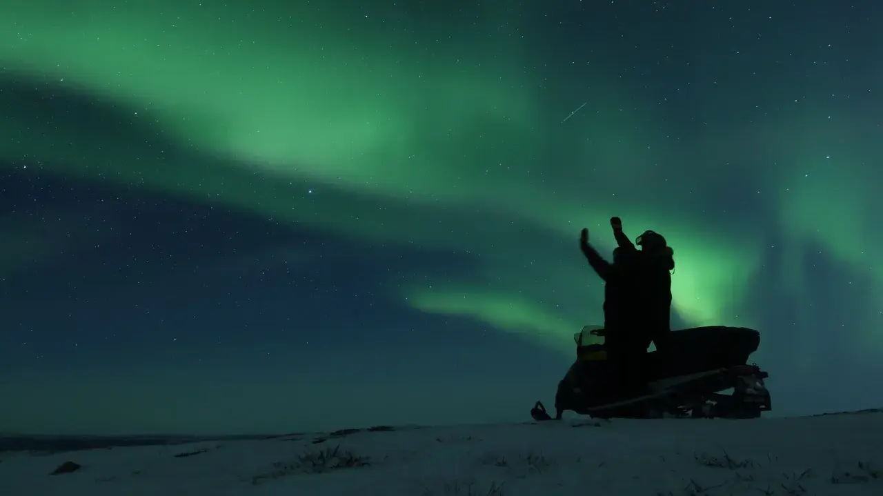 En person på snöskoter i Kiruna Lapland sträcker upp armarna mot norrskenet som lyser grönt på natthimlen.