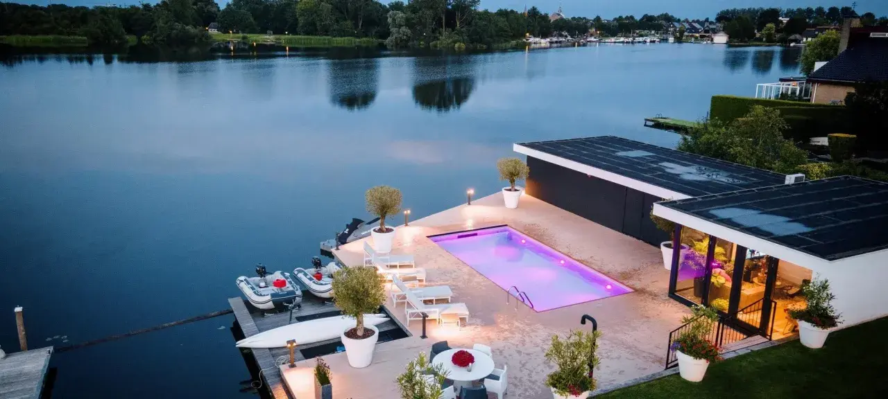 Lyxig pool med platssvetsad liner och belysning, omgiven av solstolar och växter. Båtar ligger vid bryggan.