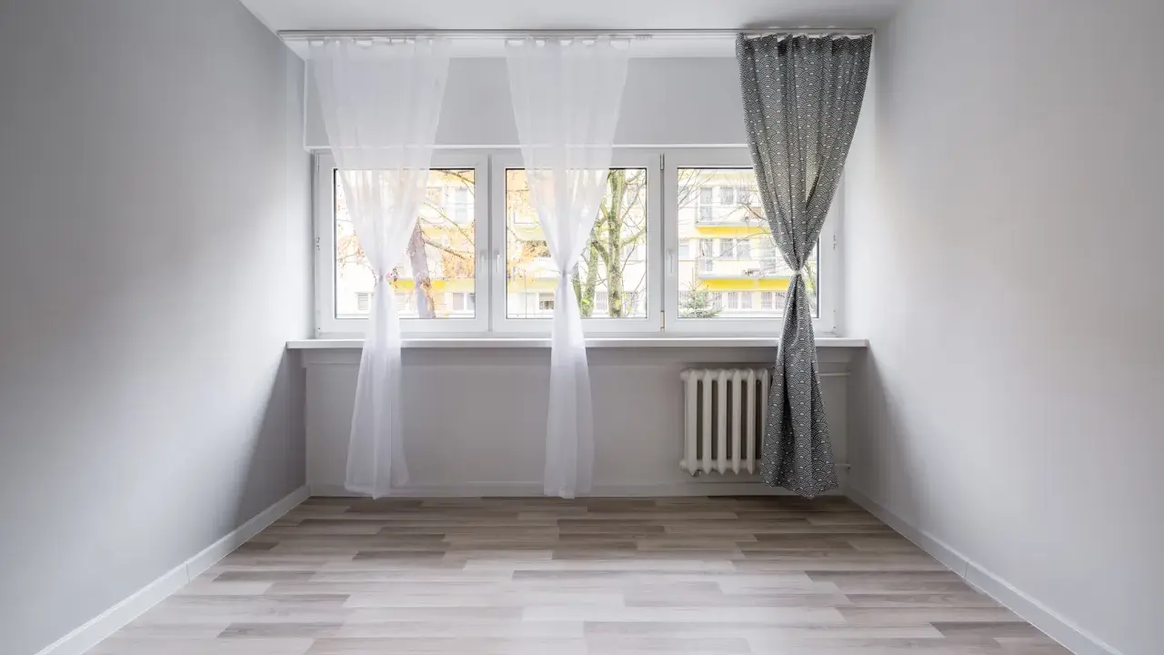 Biały karnisz do sufitu czy ściany, z białymi i wzorzystymi zasłonami, nad oknem z widokiem na budynek mieszkalny.