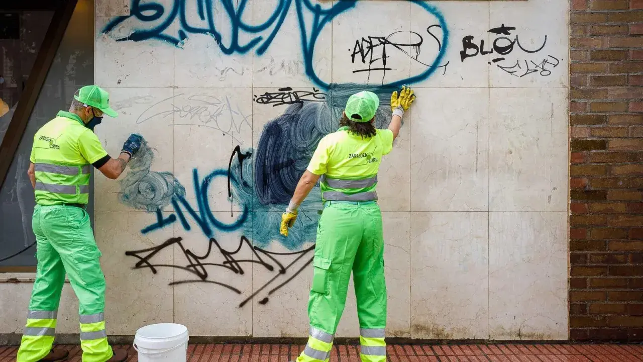 Dos trabajadores con uniformes verdes y amarillos brillantes trabajan para limpiar grafitis de una pared de mármol.