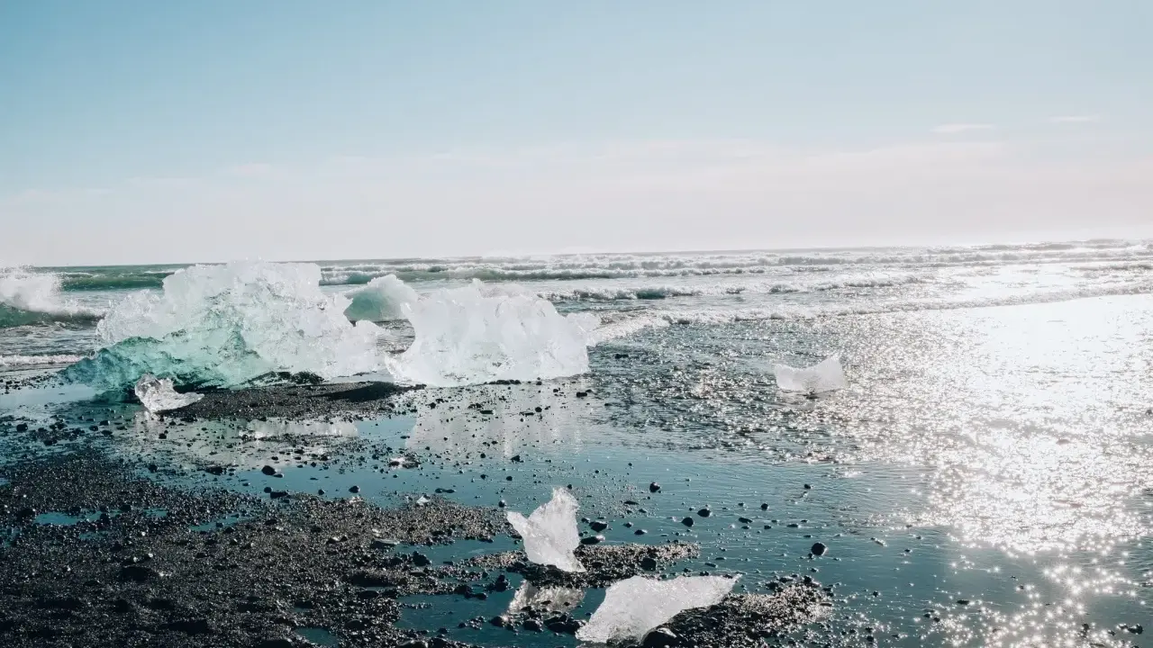 Lodowe bryły lśnią na czarnym piasku, tworząc magiczny diamond beach. Fale obmywają brzeg, a słońce odbija się w wodzie.