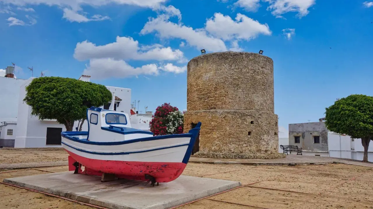 Ein Fischerboot und ein alter Turm in der conil altstadt. Weiße Häuser und grüne Bäume unter blauem Himmel mit Wolken.