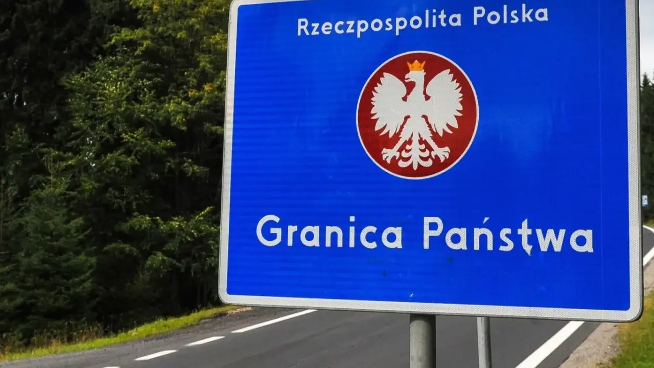 Niebieski znak z orłem i napisem "Rzeczpospolita Polska" oraz "Granica Państwa" stoi przy drodze otoczonej lasem.