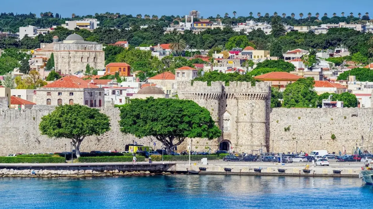 Widok na zabytkowe mury obronne i zabudowania wyspy Rodos, z błękitnym morzem w tle.