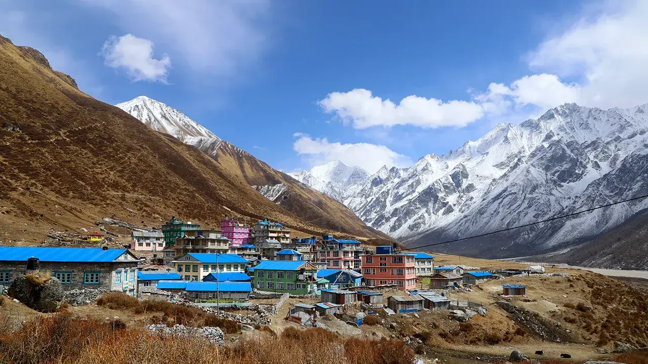 Villaggio colorato ai piedi dell'alta montagna himalayana, con case dai tetti blu e rosa, circondato da pendii bruni e cime innevate.