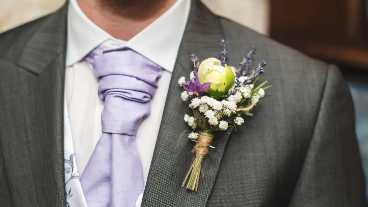 Elegancka butonierka w marynarce, ozdobiona białymi kwiatkami i lawendą, dodaje uroku ślubnemu strojowi.