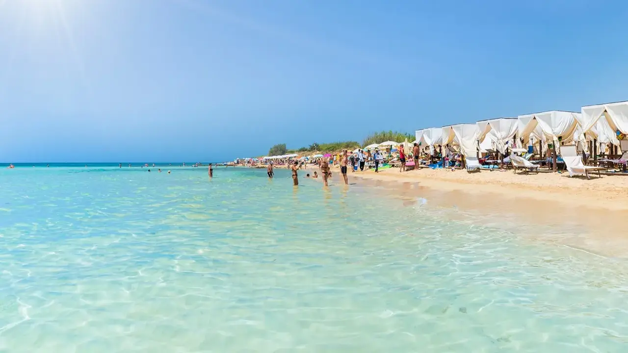 Spiaggia affollata con lettini e ombrelloni bianchi, gente che nuota e prende il sole. Un'immagine perfetta delle località balneari Puglia.