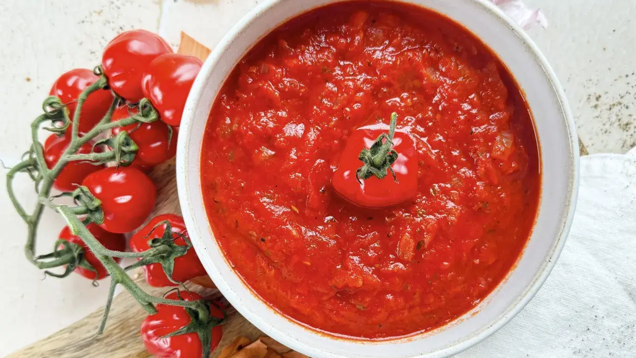 Eine Schüssel mit köstlicher, hausgemachter italienischer Tomatensauce, garniert mit einer frischen Kirschtomate. Daneben liegen frische Tomaten und Zwiebeln.