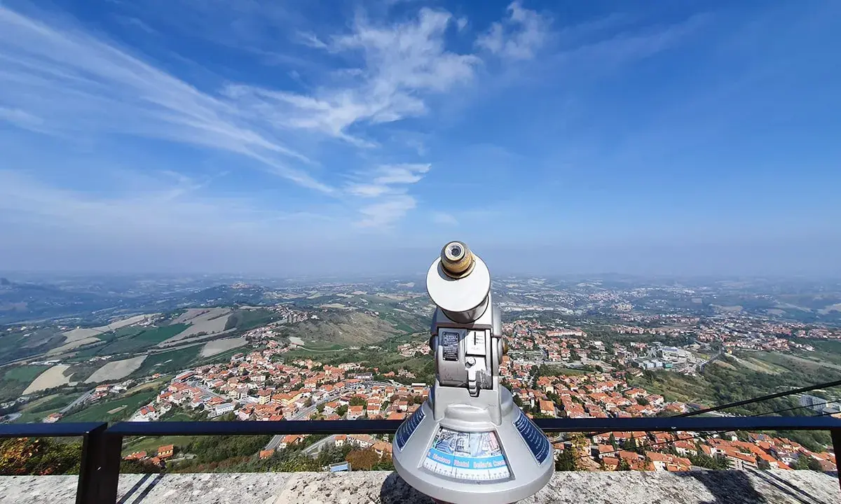 Telescopio con vista panoramica su cosa vedere a San Marino. Il paesaggio collinare e la città si estendono sotto un cielo azzurro con nuvole sottili.