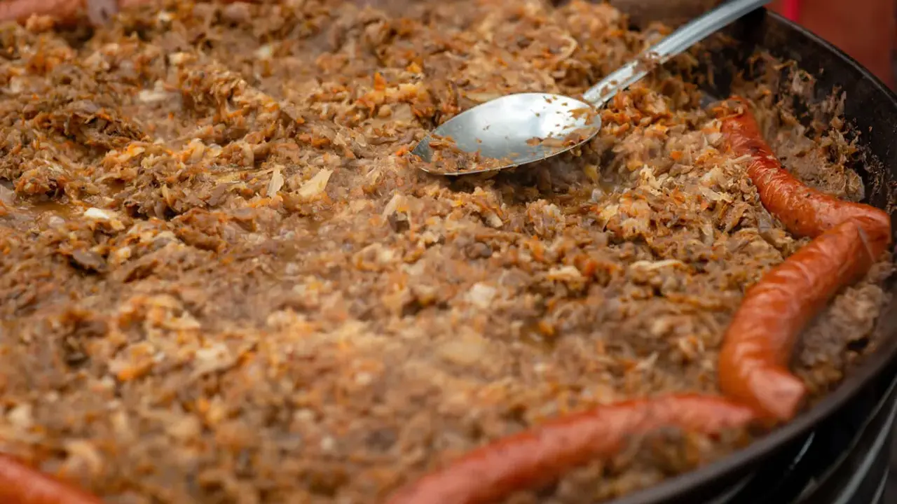 Wielki gar bigosu z kiełbasą, gotowy do wekowania w piekarniku. Łyżka zanurzona w aromatycznym daniu.
